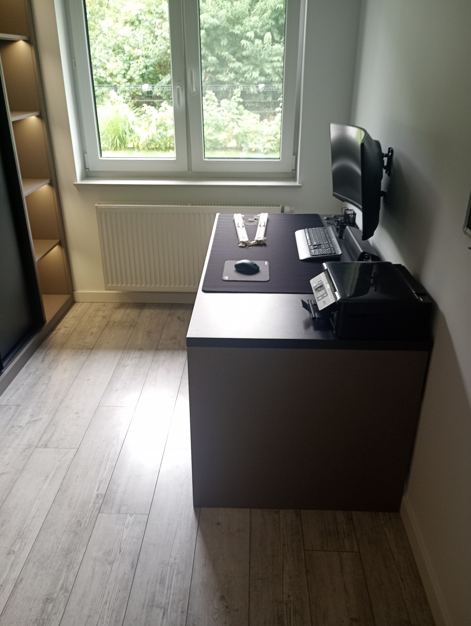 Nowoczesny pokój z biurkiem, komputerem i szafą wnękową. Minimalistyczny design, jasne panele podłogowe i duże okno z widokiem na zieleń. Szafa z oświetleniem.