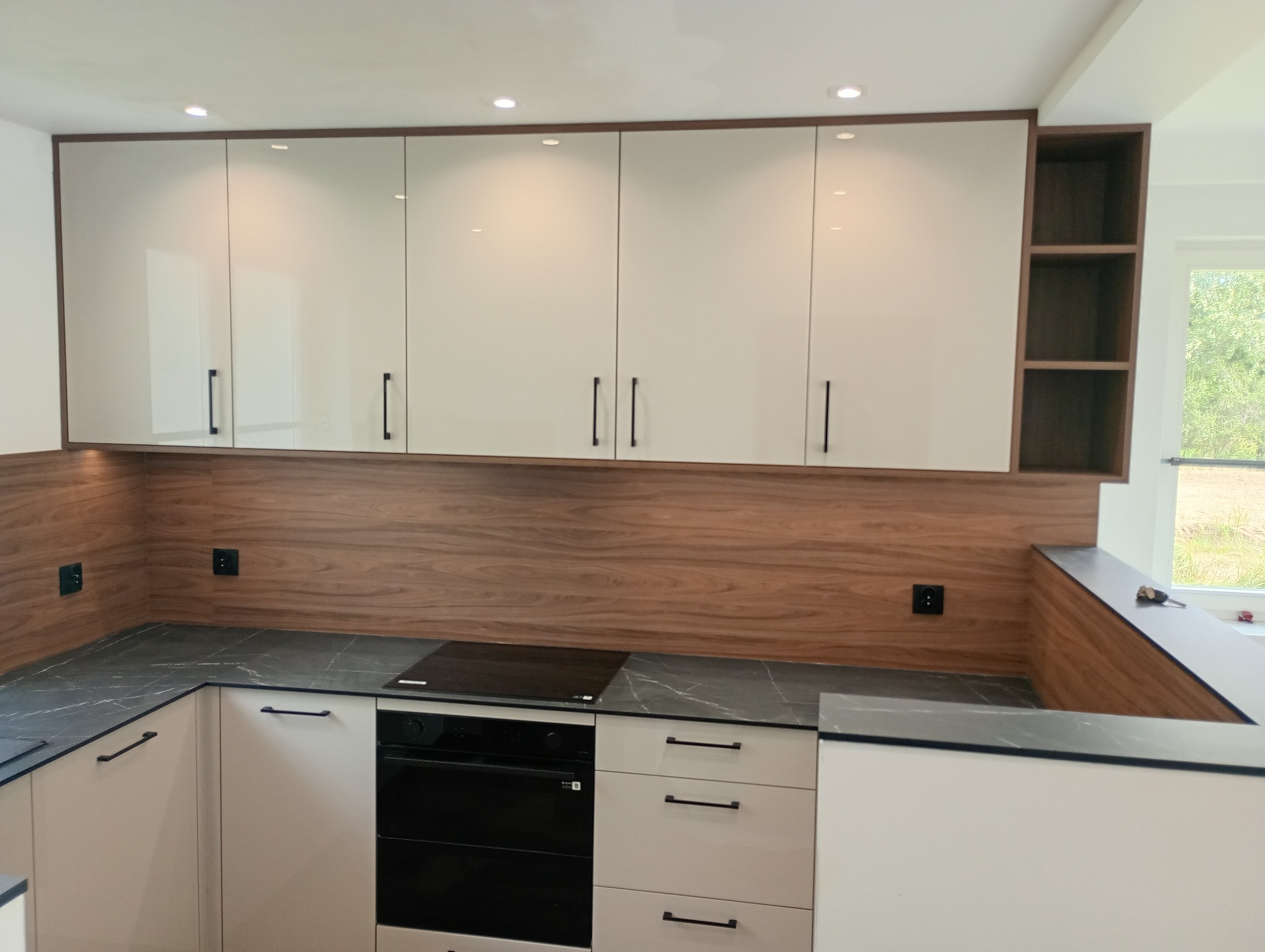 Nowoczesna kuchnia: białe szafki górne i dolne z czarnymi uchwytami, drewniany panel ścienny, czarny blat z marmurkowym wzorem i wbudowana płyta grzewcza.