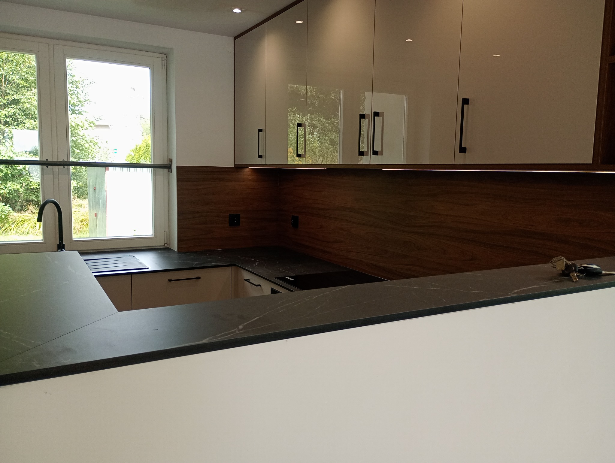 Nowoczesna kuchnia z czarnym blatem i drewnianą ścianą nad blatem, widok na okno i zlew. Na blacie leżą klucze.