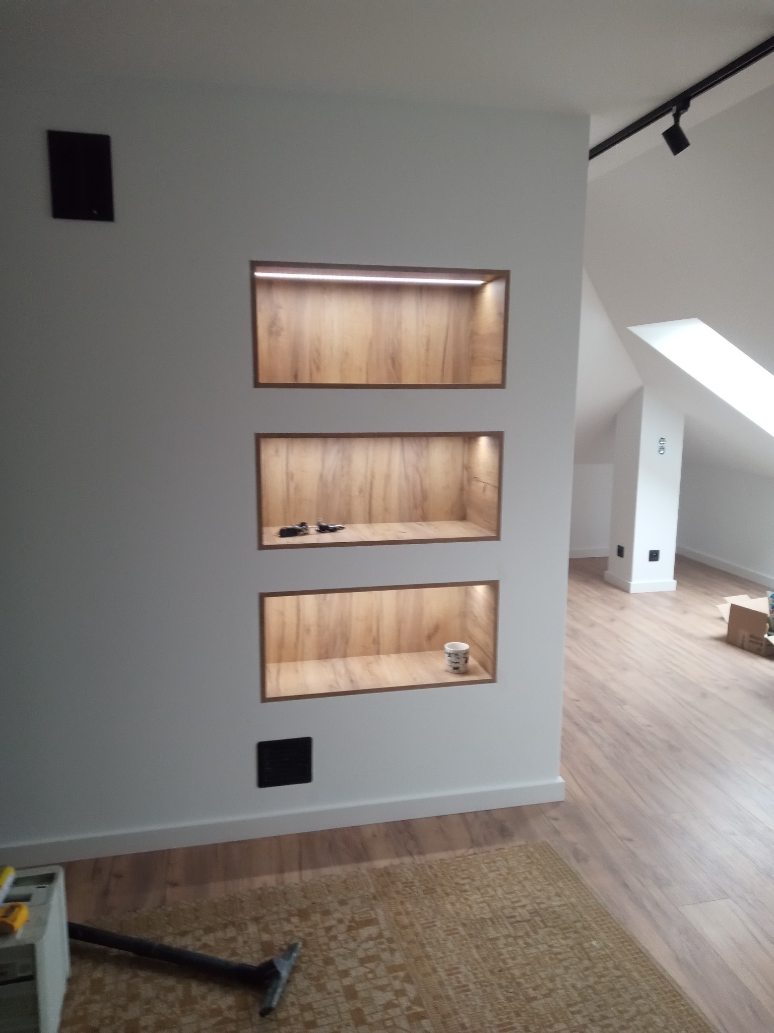 Biała ściana z trzema podświetlanymi wnękami z drewnianym wykończeniem. W dolnej wnęce stoi biały kubek. Podłoga drewniana, widoczny fragment odkurzacza.