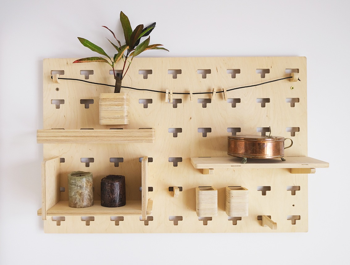 Modułowy organizer ścienny z jasnej sklejki z perforacją, na którym umieszczono małą roślinę w drewnianym wazonie, miedzianą szkatułkę na nóżkach, dwie świece oraz drewniane pojemniki, połączone...