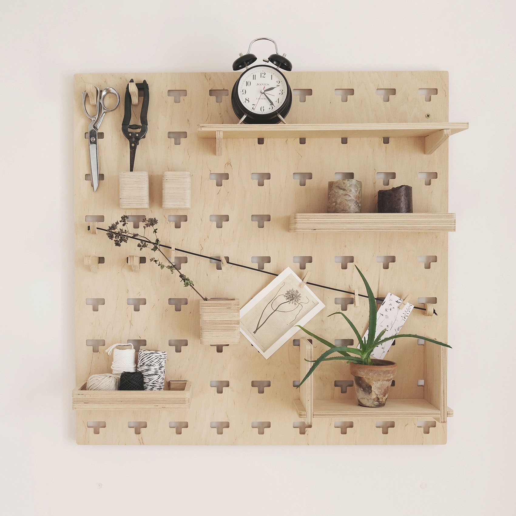 Modułowy organizer ścienny z jasnej sklejki z akcesoriami: nożyczki, sekator, szpulki nici, zegar, świece, roślina w doniczce, ilustracje, zamocowany na białej ścianie.