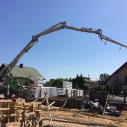 Betonomieszarka z wysięgnikiem w trakcie wylewania betonu na fundament budynku jednorodzinnego, widoczne szalunki, zbrojenia, palety z bloczkami H+H oraz grupa robotników nadzorujących pracę.