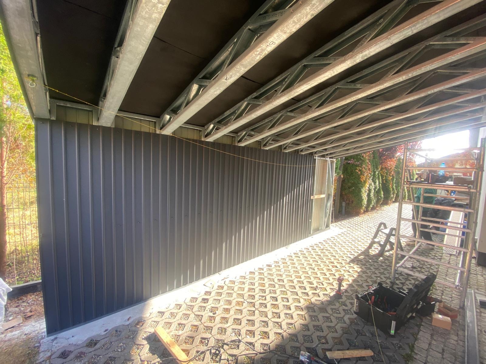 Konstrukcja wiaty z metalowym szkieletem i szarą blachą falistą, widoczny fragment dachu z izolacją, narzędzia i drabina na kostce brukowej, w tle zieleń.