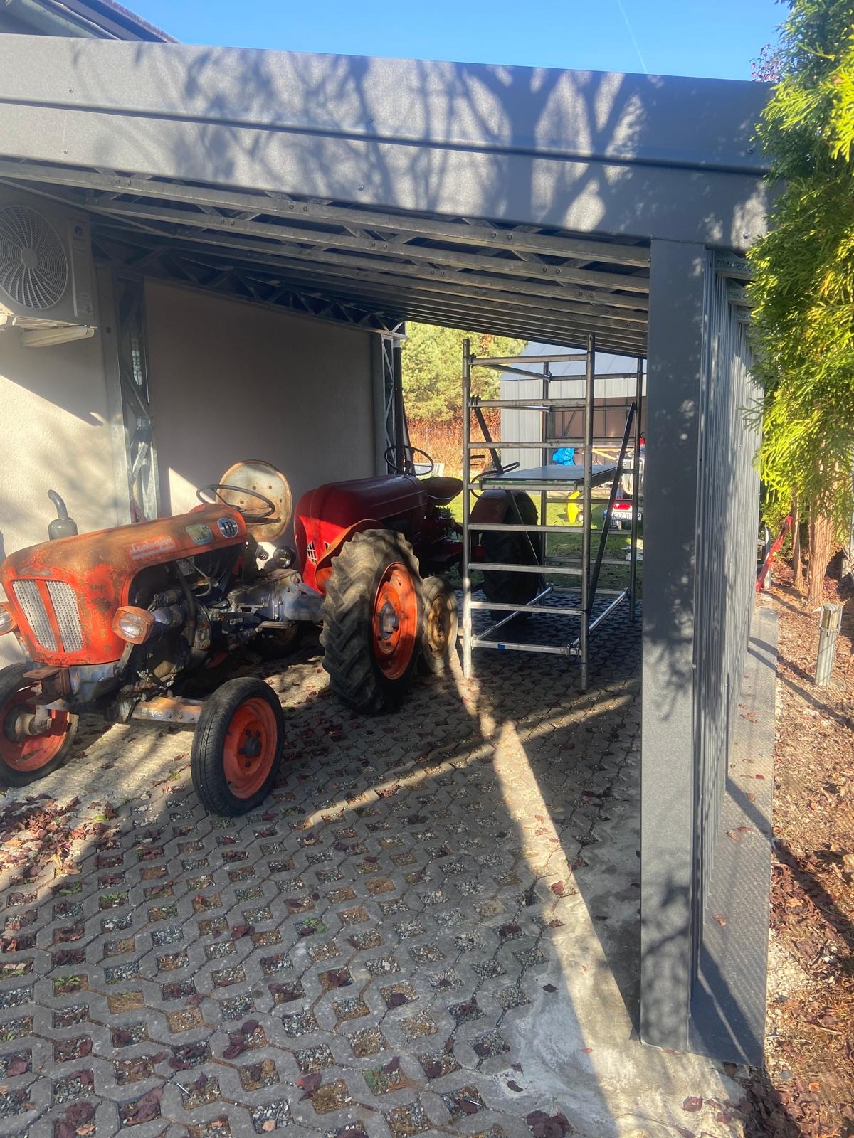 Dwa traktory, jeden z wyraźnymi śladami rdzy, stoją pod szarą, metalową konstrukcją w słoneczny dzień. Na drugim planie widoczne rusztowanie.