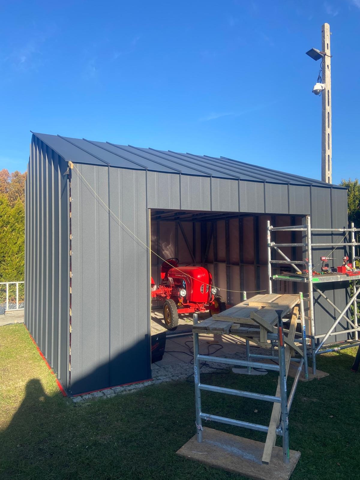 Montaż metalowej hali w Morawicy, widoczny czerwony traktor wewnątrz, rusztowania i narzędzia do montażu na zewnątrz, słoneczny dzień.
