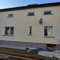 Dwukondygnacyjny dom z beżową elewacją po renowacji, widoczna antena satelitarna, nowe okna z białymi ramami i częściowo odsłonięte drewniane elementy konstrukcyjne na pierwszym planie.