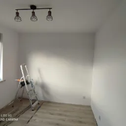 Świeżo pomalowane na biało wnętrze pokoju z drewnopodobną podłogą, widoczna aluminiowa drabina z narzędziami oraz lampa sufitowa z trzema metalowymi kloszami w stylu industrialnym.