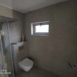 Wykończona łazienka z kabiną prysznicową, podwieszanym WC i oknem, widoczne niedociągnięcia w postaci wystających kabli i resztek po remoncie.