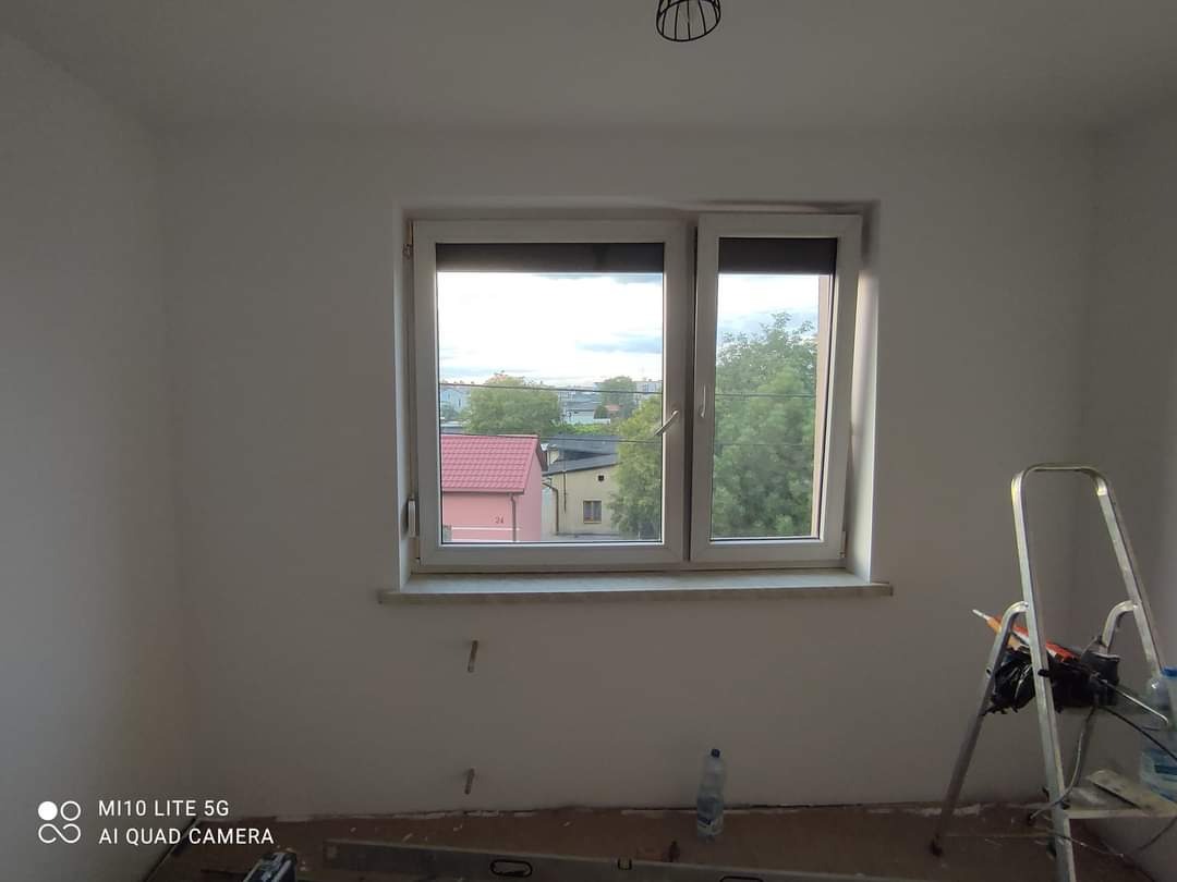 Pokój po malowaniu z widocznym oknem, drabina malarska i butelka wody na podłodze, ściany w kolorze białym, widoczne niedoskonałości na podłodze.