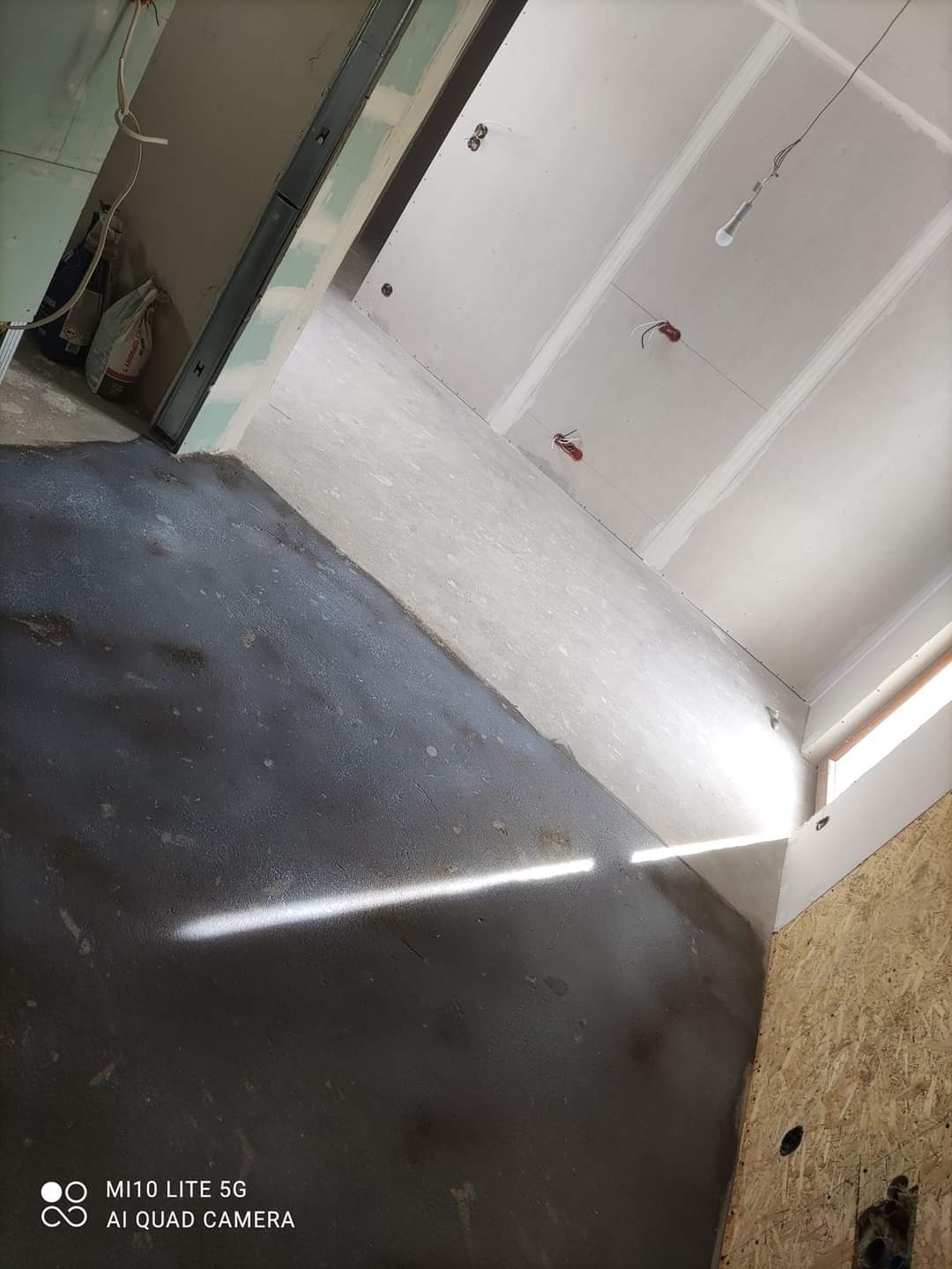 Wnętrze w trakcie prac wykończeniowych: surowy, ciemny podkład na podłodze kontrastuje z jasnymi, przygotowanymi do malowania ścianami z płyt gipsowo-kartonowych i sufitem, widoczne przewody...