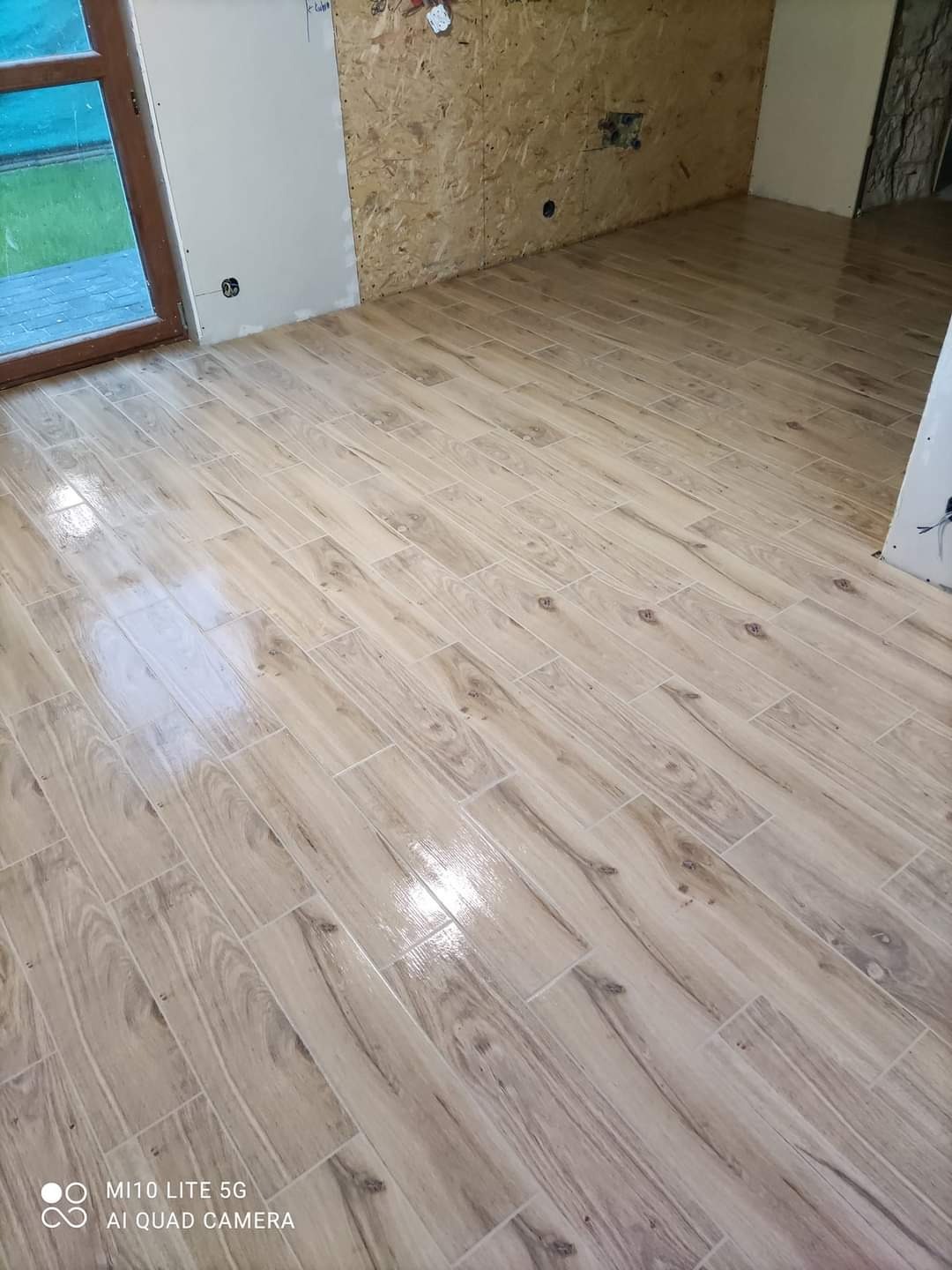 Ułożona podłoga z płytek imitujących drewno w jasnym odcieniu, widoczne surowe ściany z płyt OSB i fragment okna z widokiem na zieleń.