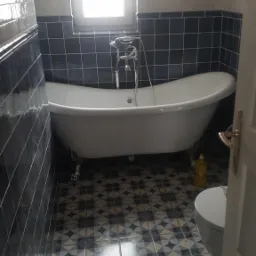 Biała wanna wolnostojąca na lwich nóżkach w łazience z ciemnymi płytkami i wzorzystą podłogą, widoczna butelka płynu do czyszczenia.