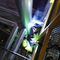 Spaw System Weldnig Group - Prace spawalnicze na wysokości: spawanie rur przemysłowych z użyciem drabiny, widoczne iskry i dym, pracownik w ochronnym ubraniu i masce.