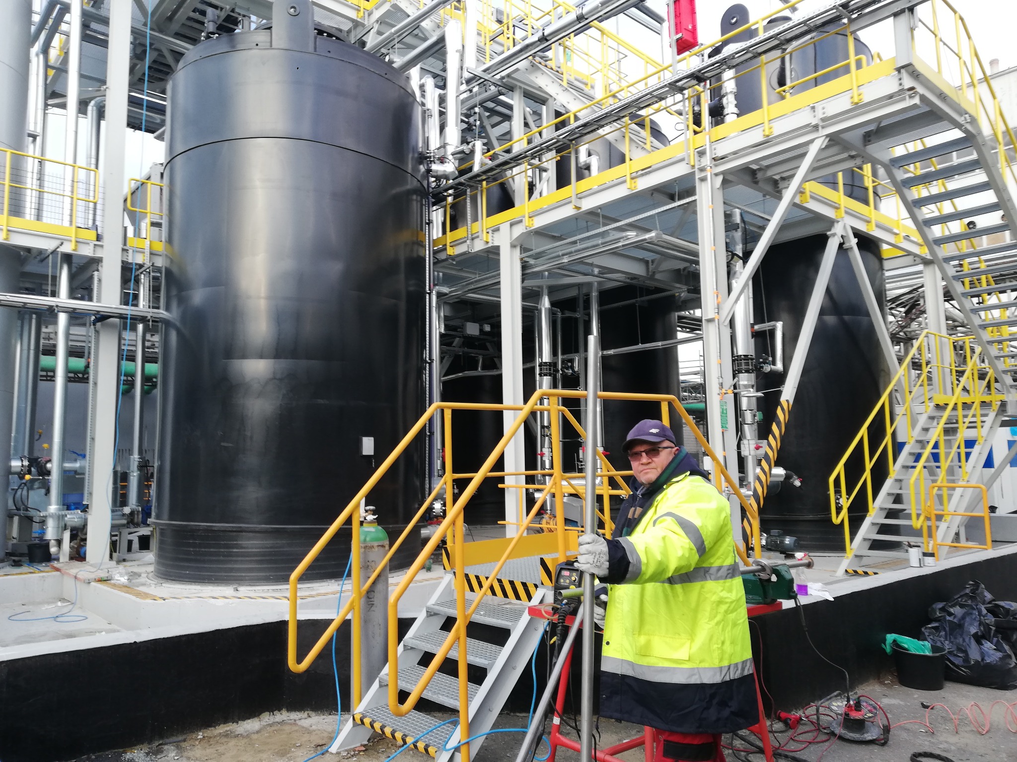 Pracownik w żółtej kurtce przy metalowej konstrukcji z dużymi zbiornikami, prawdopodobnie podczas prac montażowych lub kontrolnych.