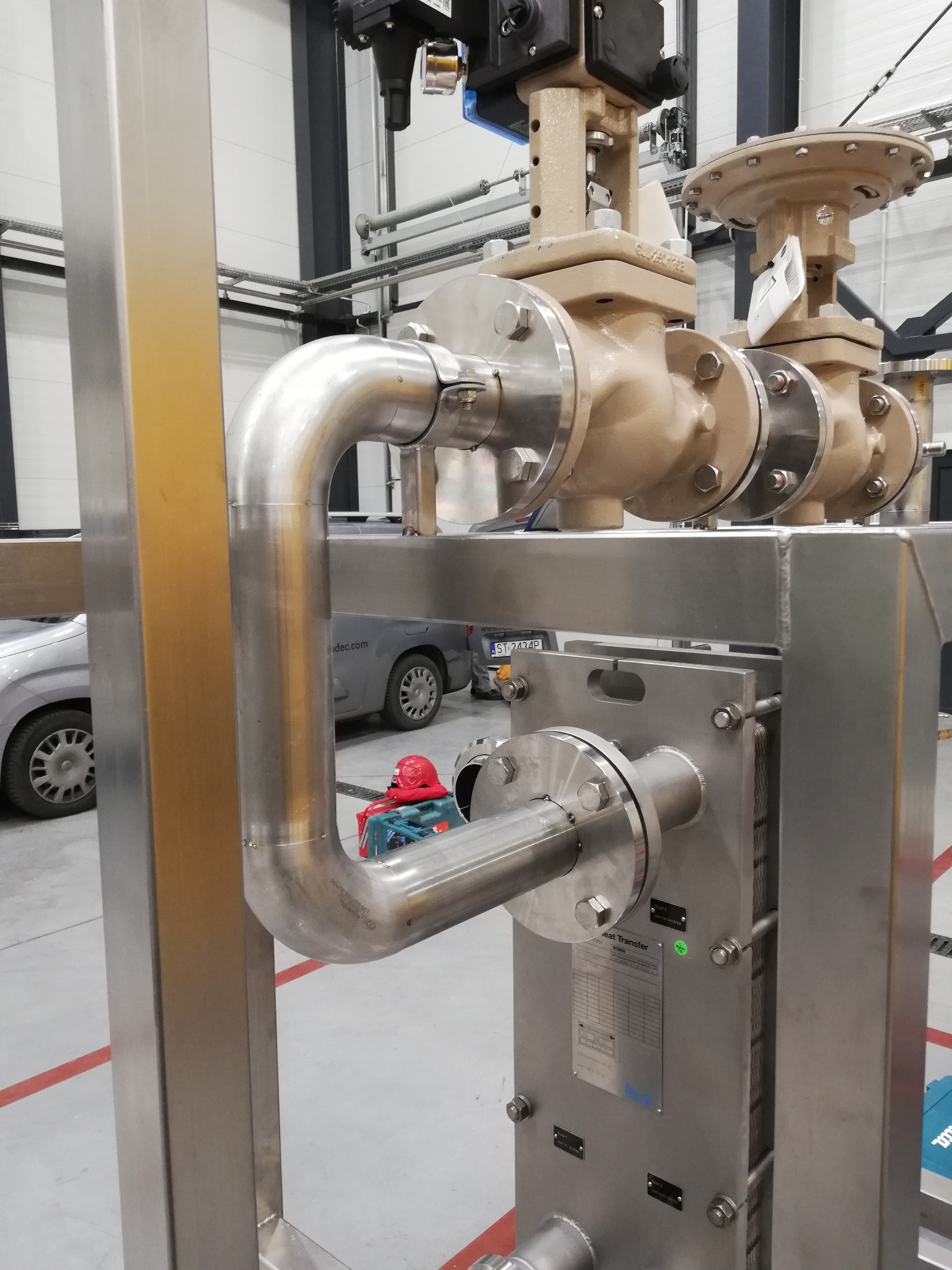 Instalacja przemysłowa ze stali nierdzewnej z zaworami i wymiennikiem ciepła, widoczny fragment samochodu i narzędzia w tle.