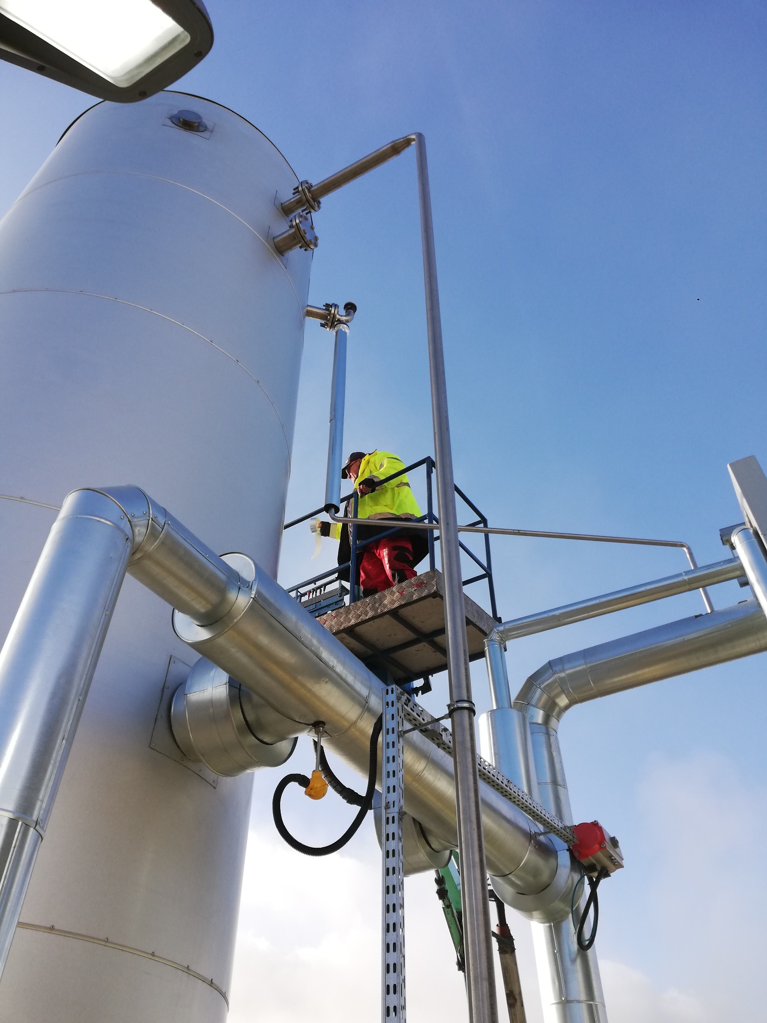 Pracownik w żółtej kamizelce na podnośniku montuje rury przy srebrnym zbiorniku ciśnieniowym na tle błękitnego nieba.