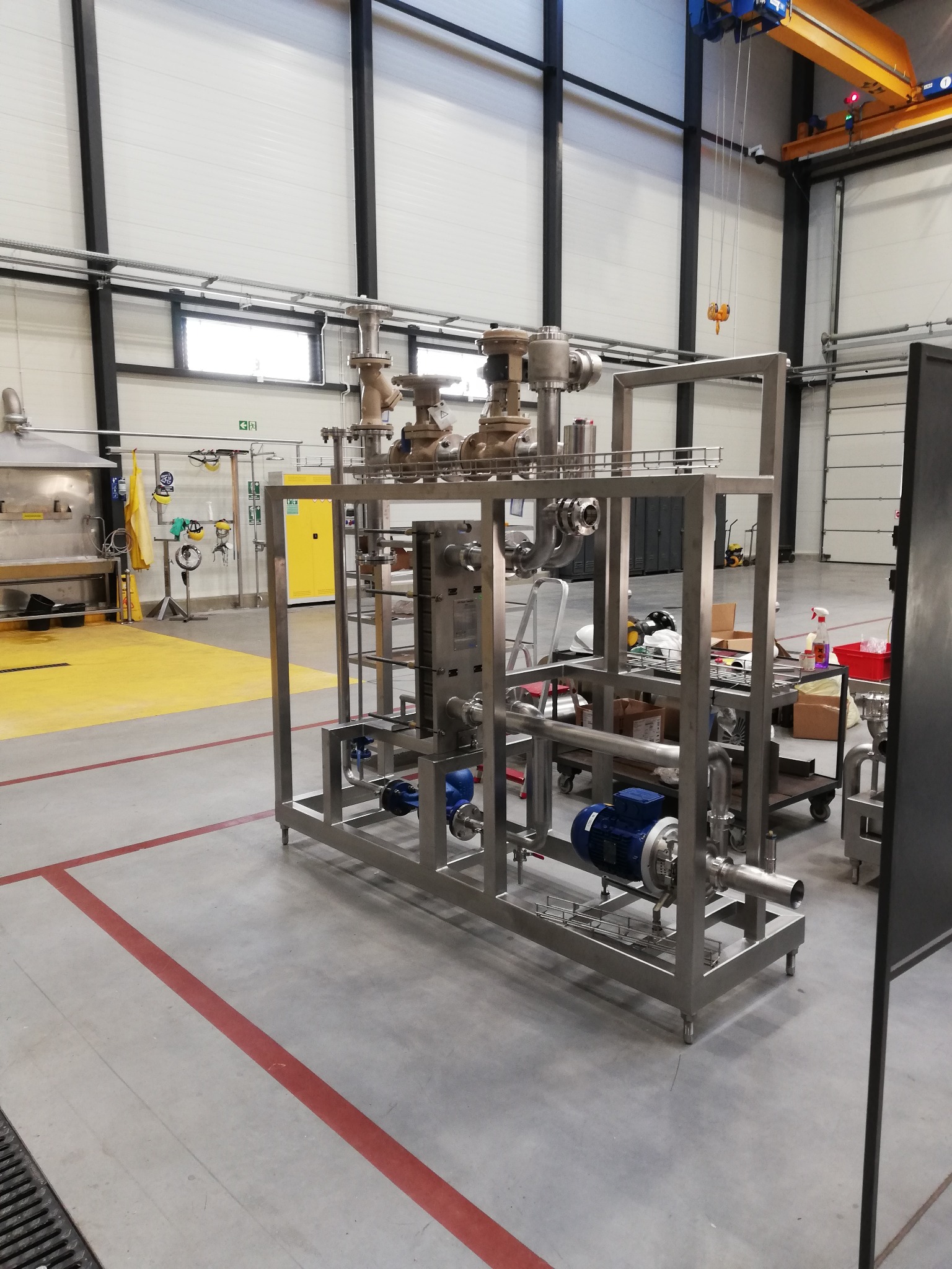 Konstrukcja stalowa z rurami, zaworami i silnikiem elektrycznym w hali produkcyjnej z suwnicą pod stropem.