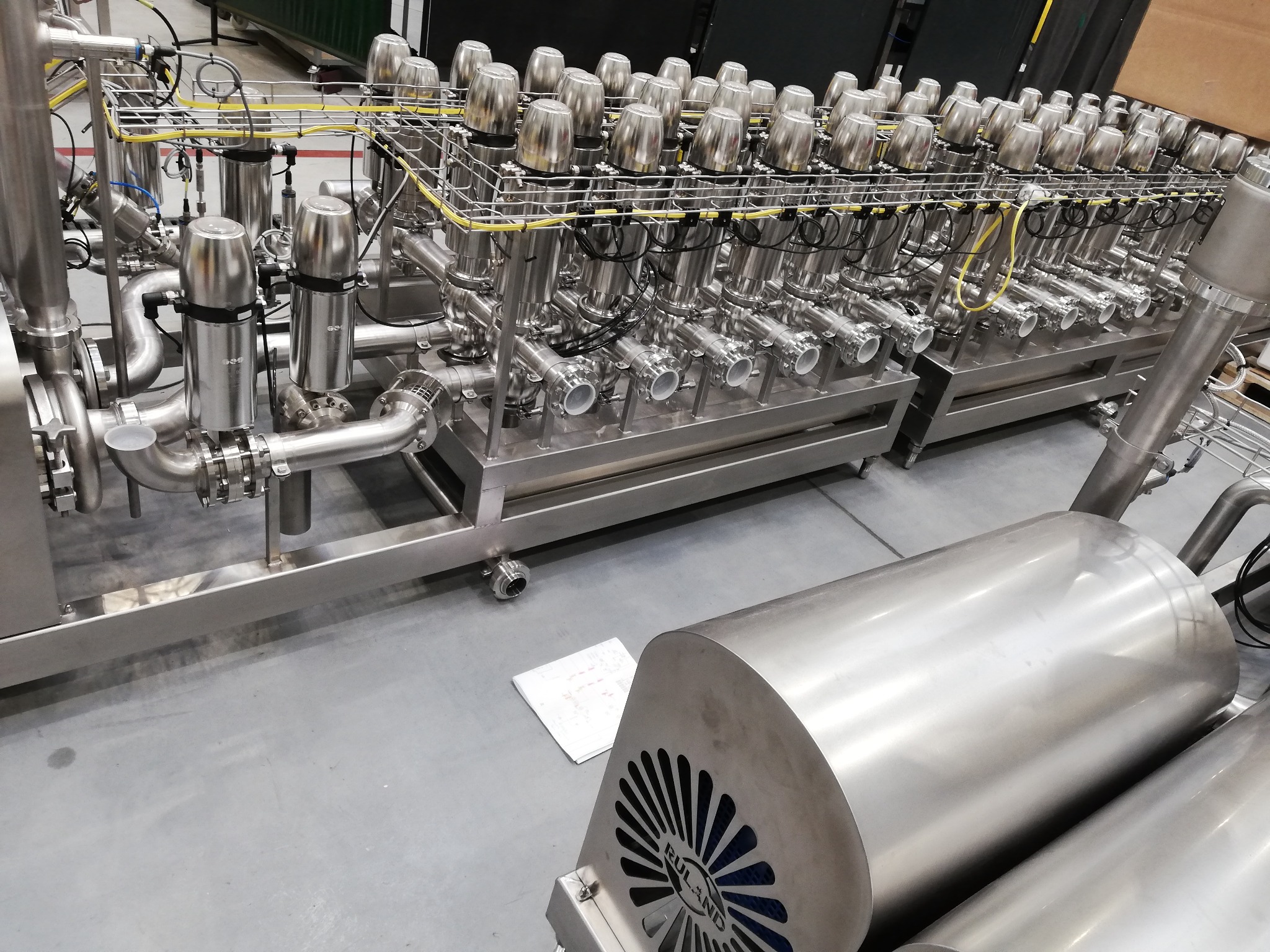 Kompleksowa instalacja przemysłowa ze stali nierdzewnej z licznymi zaworami, rurami i przewodami, widoczne elementy automatyki i dokumentacja techniczna obok.