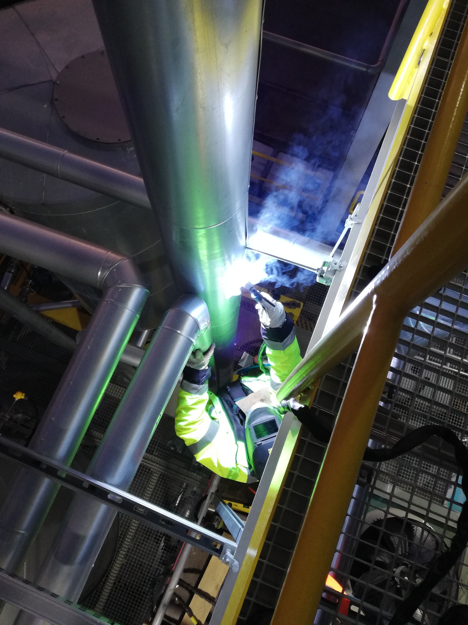 Prace spawalnicze na wysokości: spawanie rur przemysłowych z użyciem drabiny, widoczne iskry i dym, pracownik w ochronnym ubraniu i masce.