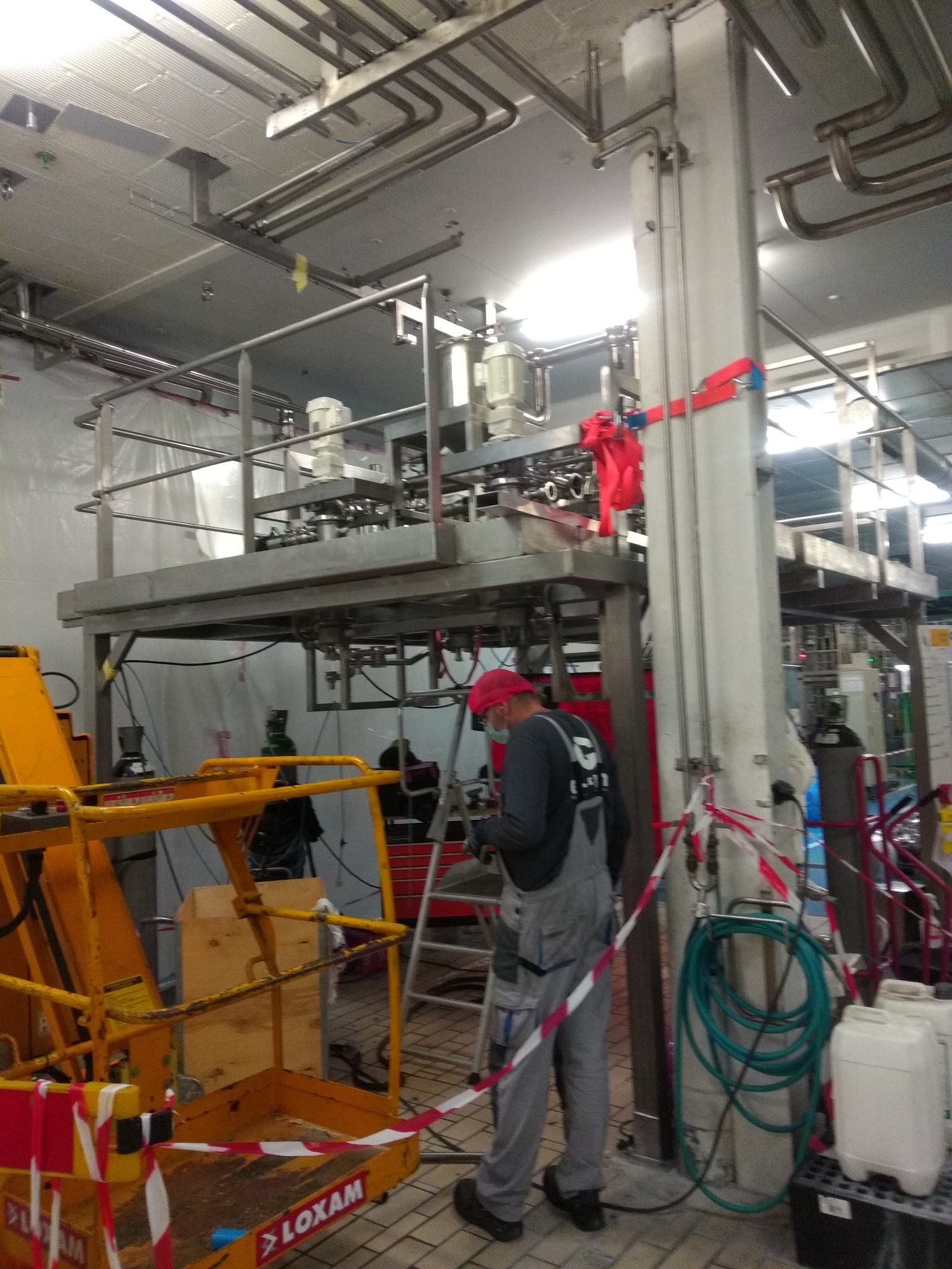 Wnętrze hali produkcyjnej z instalacją przemysłową na metalowych konstrukcjach, pracownik w kombinezonie i czerwonej czapce na drabinie, żółty podnośnik LOXAM i zabezpieczenia taśmami ostrzegawczymi.
