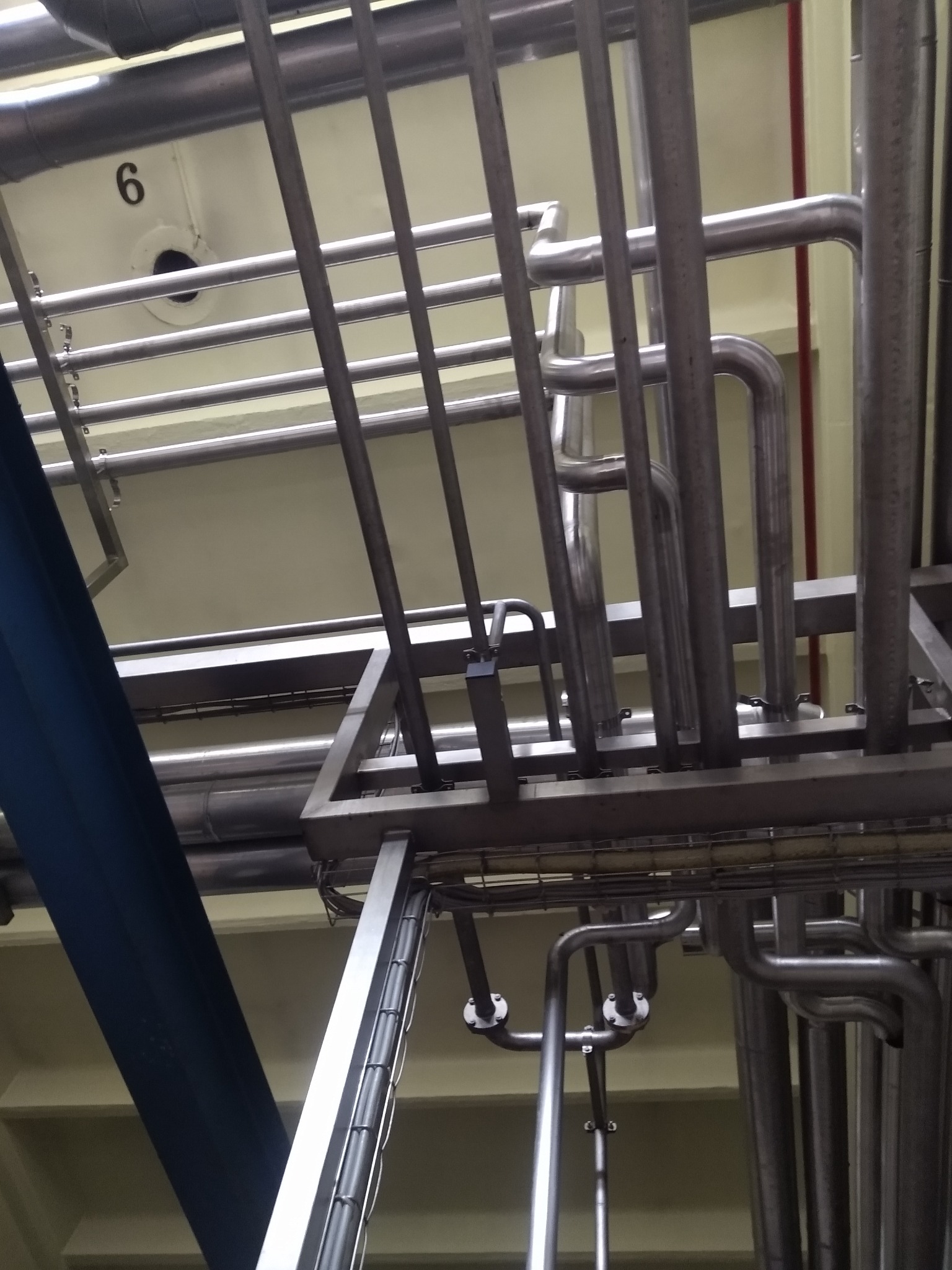 Skomplikowany układ rur ze stali nierdzewnej, widoczne liczne złącza kołnierzowe, fragment konstrukcji wsporczej w tle, numer '6' na ścianie.