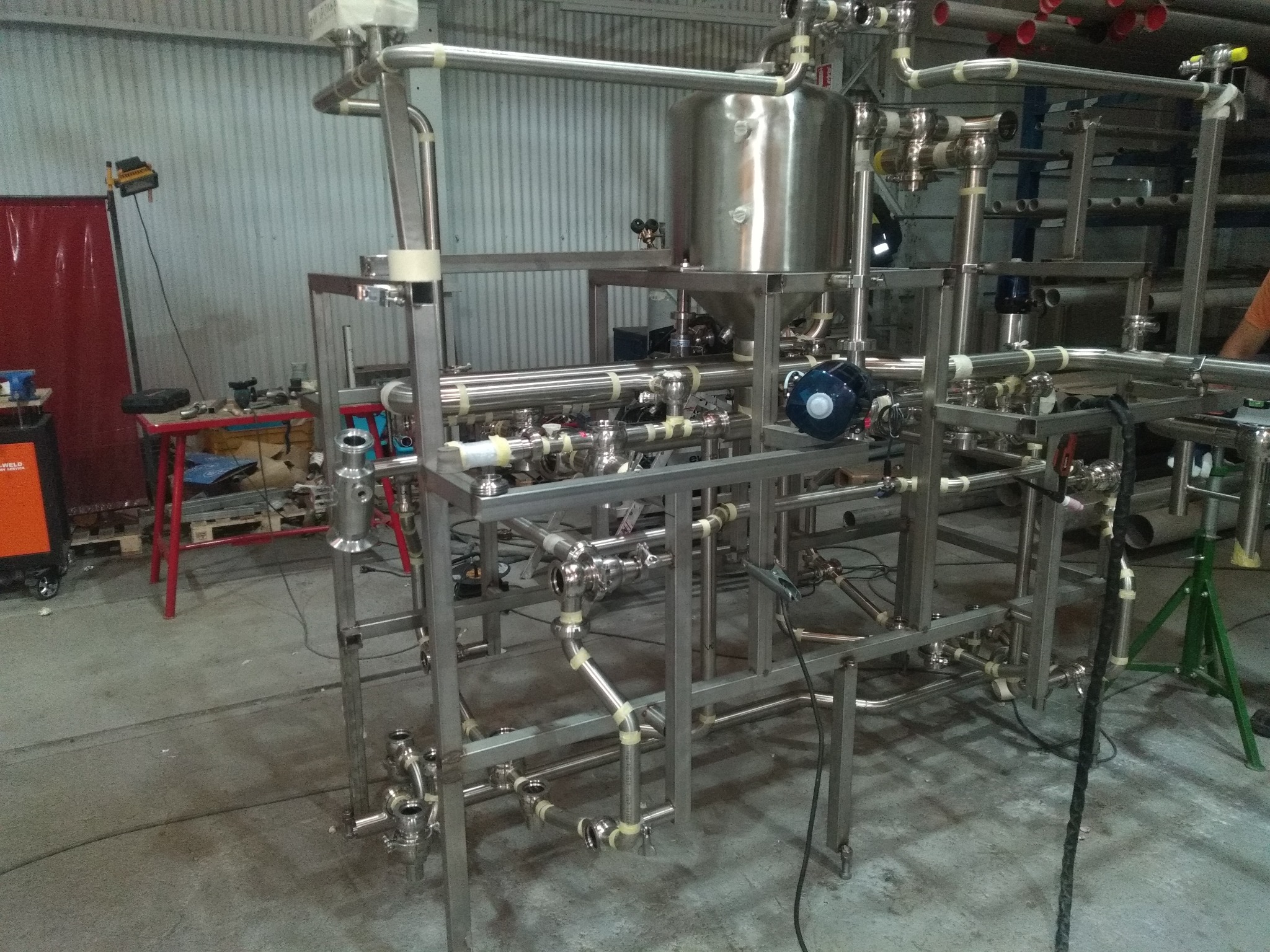 Skomplikowana instalacja rurowa ze stali nierdzewnej z zaworami i zbiornikiem, widoczna w hali produkcyjnej, z elementami tymczasowo zabezpieczonymi taśmą, spawarka i stół warsztatowy w tle.