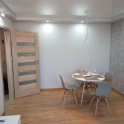 Jasne wnętrze mieszkania po remoncie: drewniana podłoga, okrągły stół z czterema krzesłami, dekoracyjna ściana z geometrycznym wzorem oraz ściana z trójwymiarową strukturą. Widoczna również...