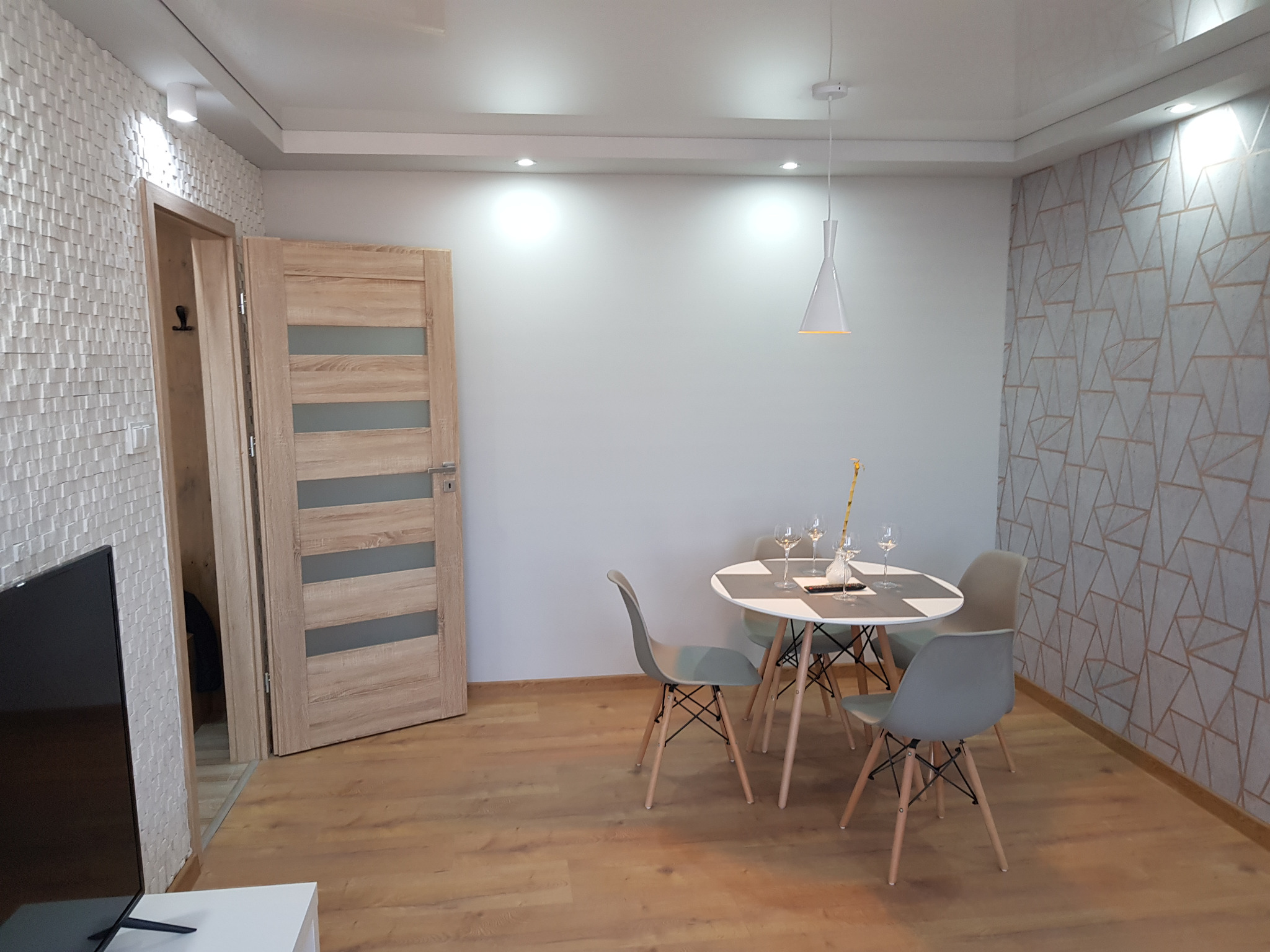 Jasne wnętrze mieszkania po remoncie: drewniana podłoga, okrągły stół z czterema krzesłami, dekoracyjna ściana z geometrycznym wzorem oraz ściana z trójwymiarową strukturą. Widoczna również...