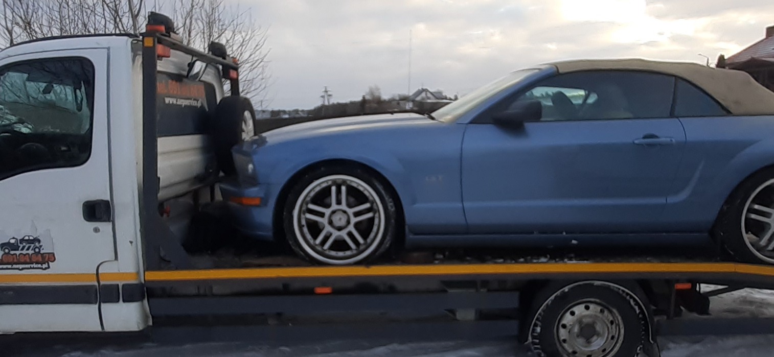 Niebieski kabriolet Ford Mustang na platformie lawety w zimowych warunkach; widoczny numer telefonu i adres strony internetowej firmy na kabinie lawety.