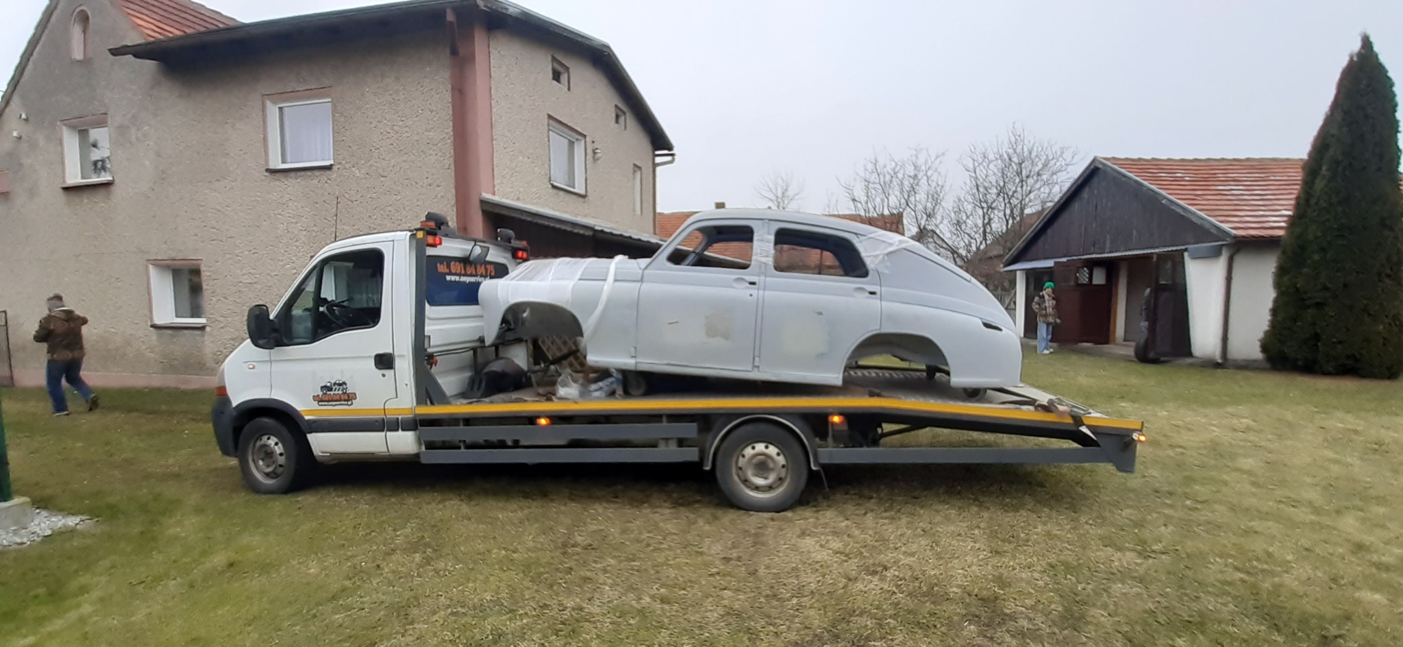 Laweta przewozi karoserię klasycznego samochodu marki Warszawa M20, zabezpieczoną folią, na tle wiejskiego krajobrazu z domami.