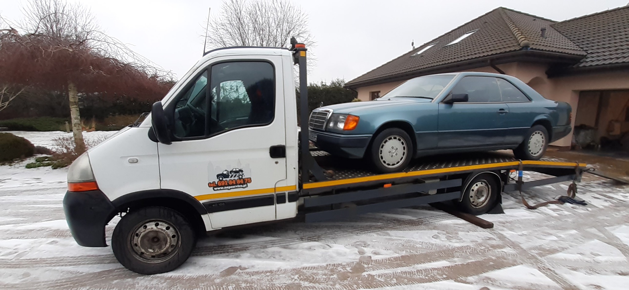 Laweta z załadowanym niebieskim Mercedesem Coupe na tle domu w zimowej scenerii.