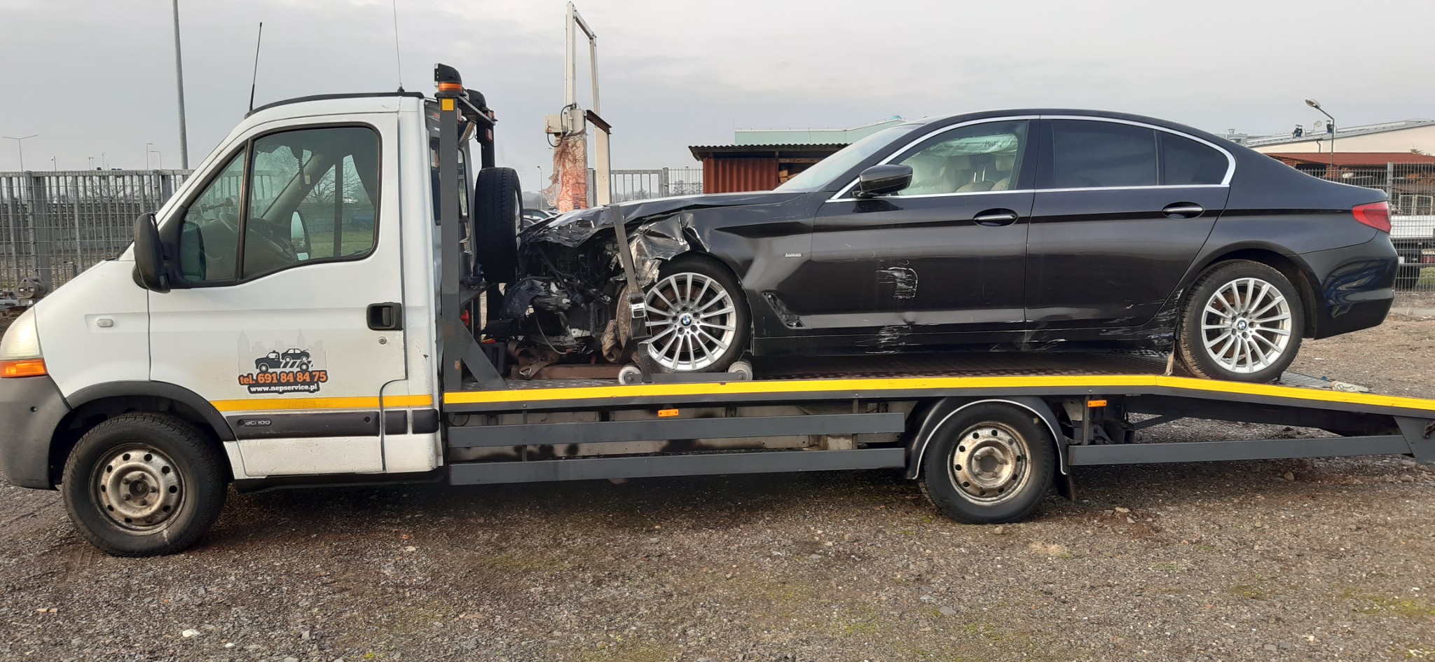 Czarny samochód BMW z uszkodzonym przodem załadowany na platformę lawety, widoczny z boku, z logiem firmy na białej kabinie lawety i zapasowym kołem z przodu.