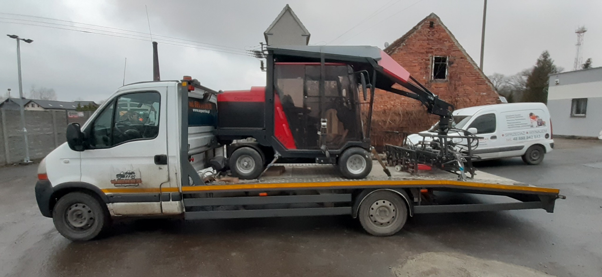 Laweta z załadowanym specjalistycznym traktorem do pielęgnacji trawy na tle ceglanego budynku i zaparkowanego białego busa.