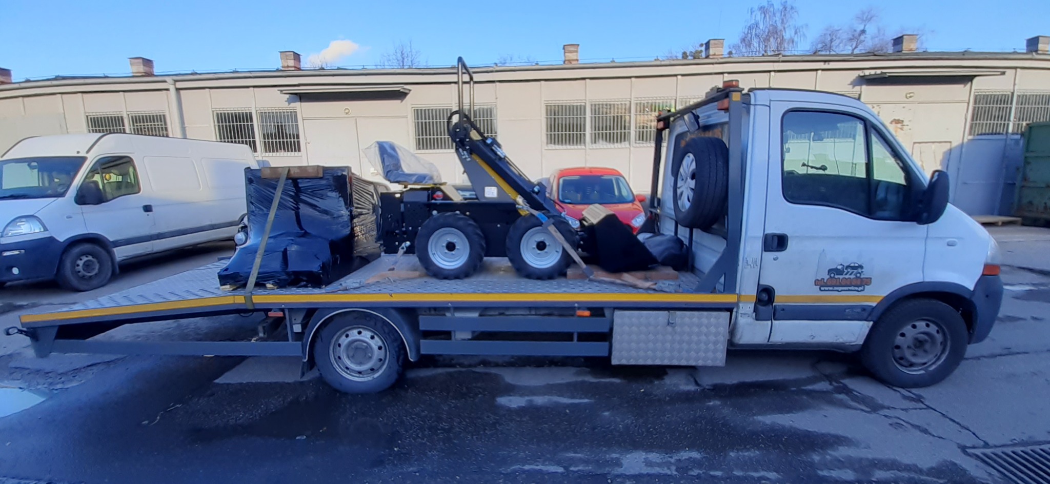 Laweta z miniładowarką i zapasowym kołem, zaparkowana na tle budynku magazynowego, obok zaparkowany biały bus.