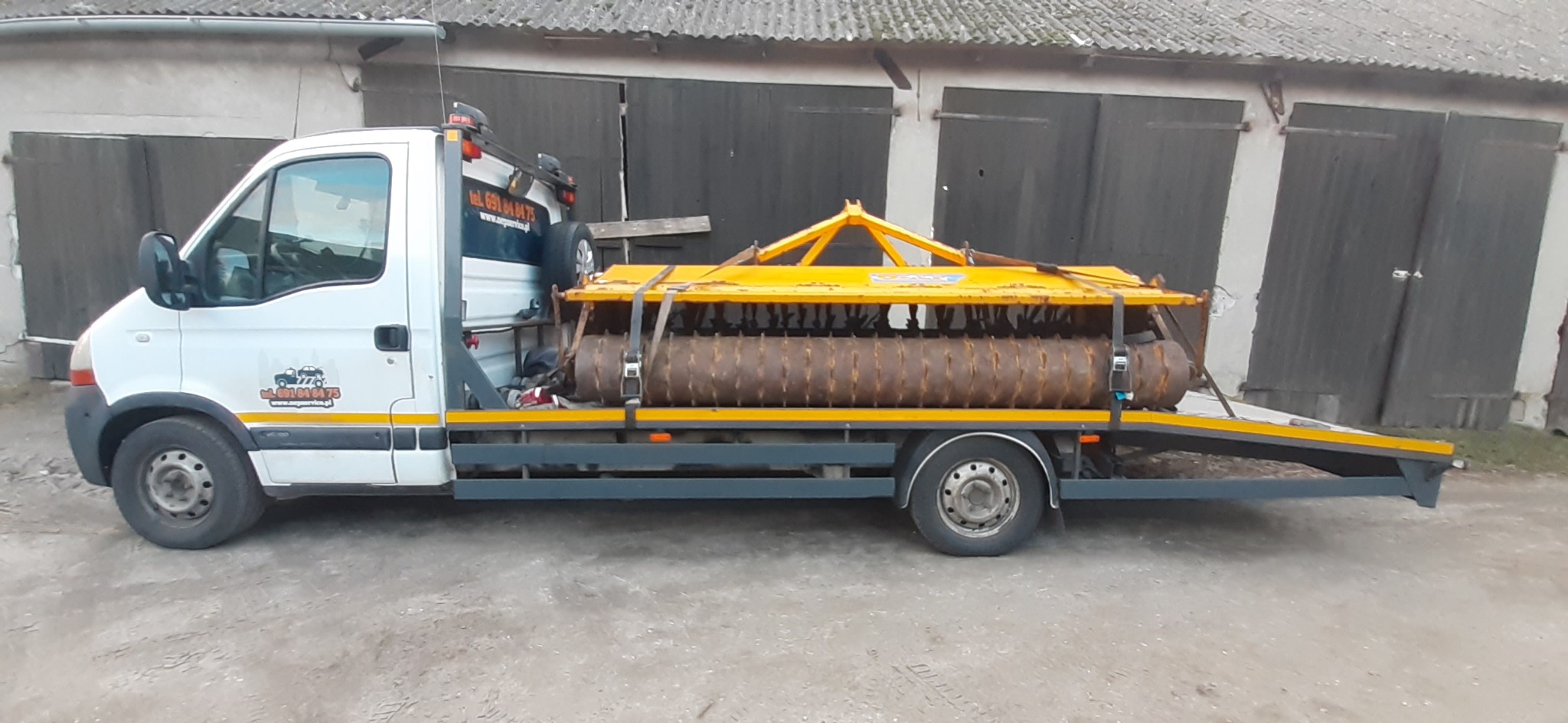 Biała laweta z żółto-brązowym wałem rolniczym na platformie, zaparkowana przed budynkiem gospodarczym z szarymi wrotami.