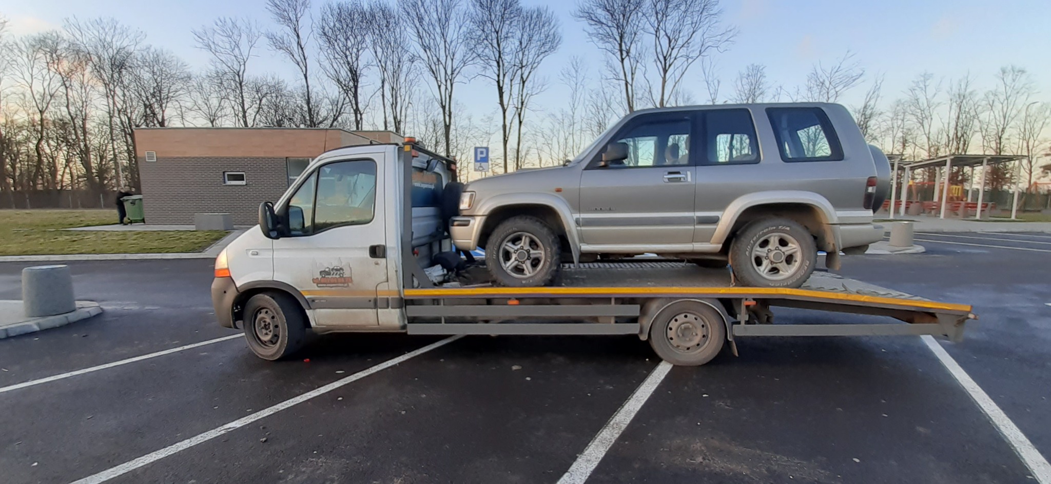 Srebrny samochód osobowy na platformie lawety, widoczny znak P dla parkingu w tle, dzień.