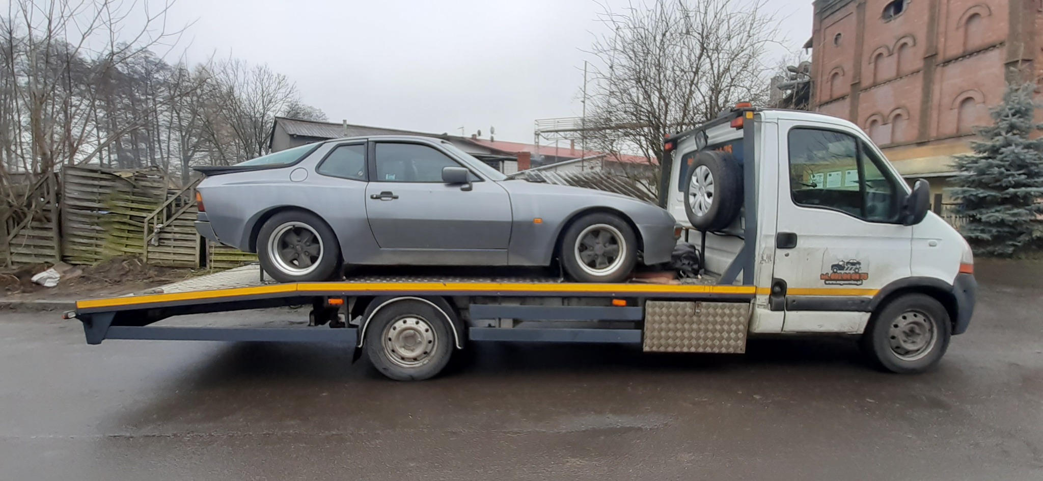 Szary samochód sportowy Porsche 944 na platformie lawety, widok z boku, w tle budynek z czerwonej cegły i drzewa bez liści.