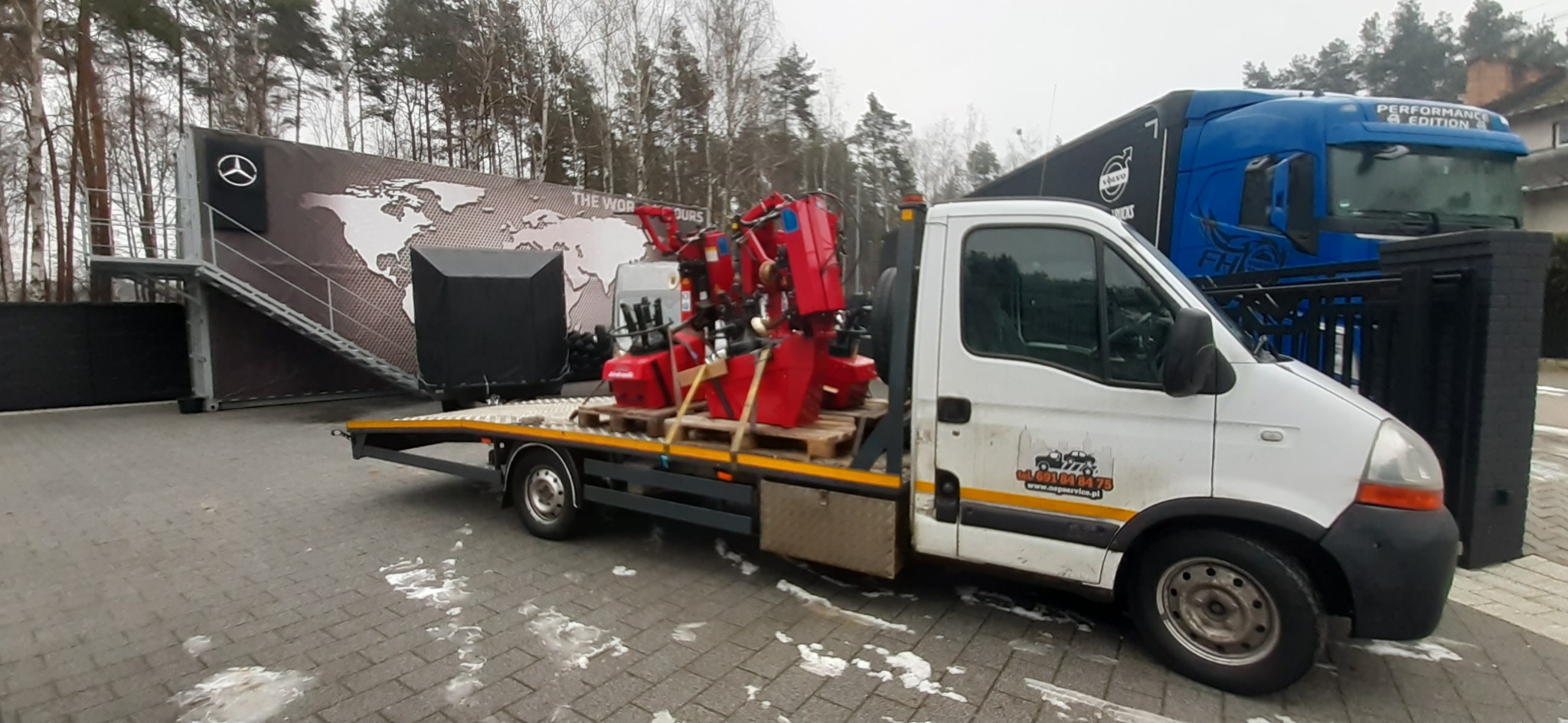 Biała laweta z czerwonymi maszynami na paletach, zaparkowana na zewnątrz, z kontenerem Mercedes-Benz w tle i niebieską ciężarówką Volvo po prawej stronie.