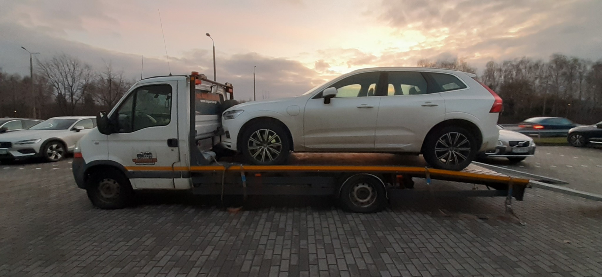 Biała laweta z załadowanym białym SUV-em Volvo na parkingu, widoczne inne zaparkowane samochody i zachodzące słońce w tle.