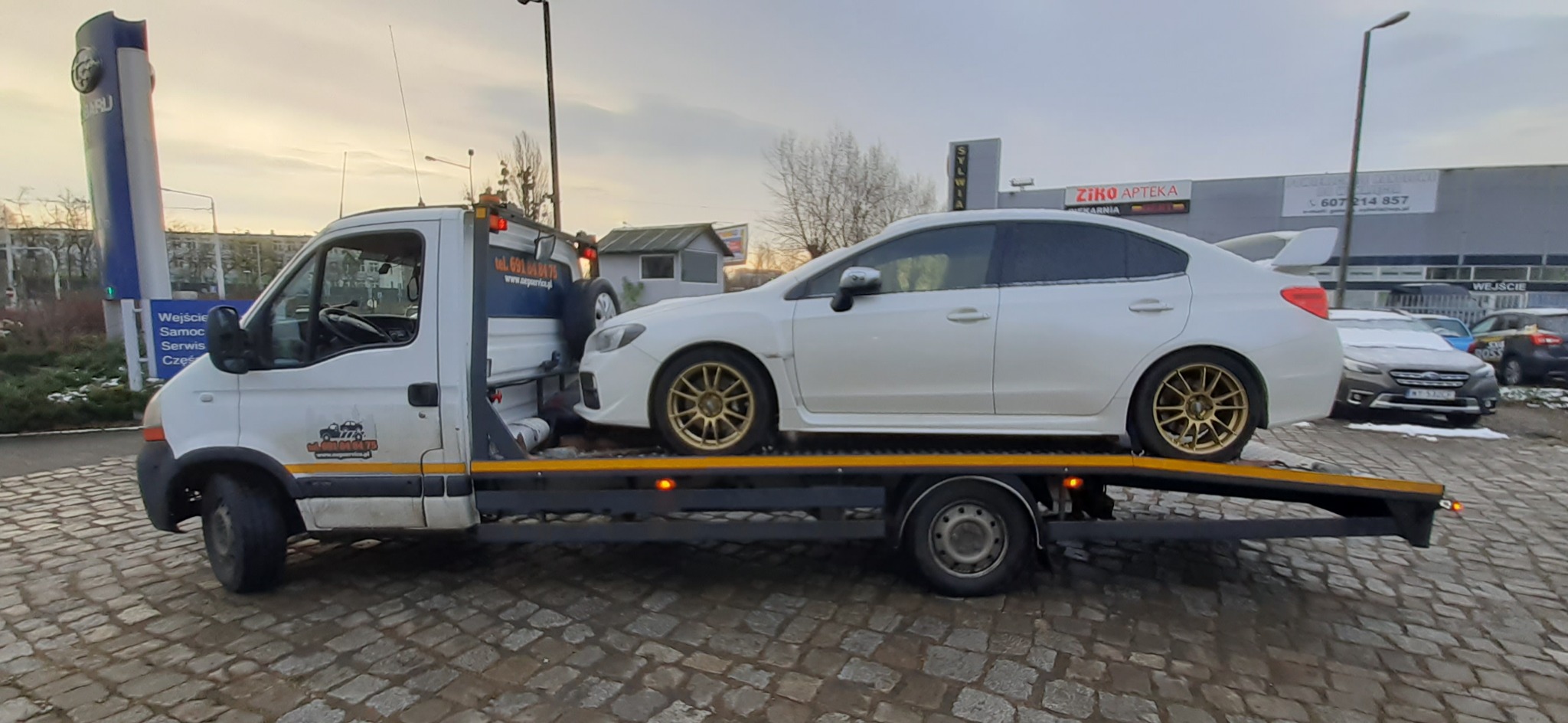 Biały samochód Subaru z złotymi felgami transportowany na lawecie na tle budynku z szyldem Ziko Apteka i salonu Subaru.