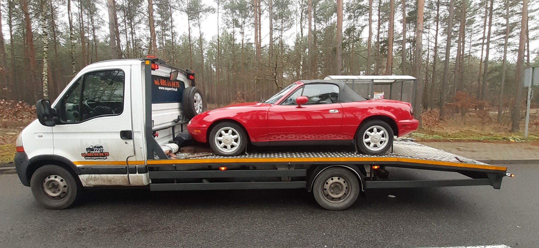 Czerwony kabriolet Mazda Miata przewożony na platformie lawety, widoczny pracownik zabezpieczający ładunek, w tle las iglasty i przystanek autobusowy.