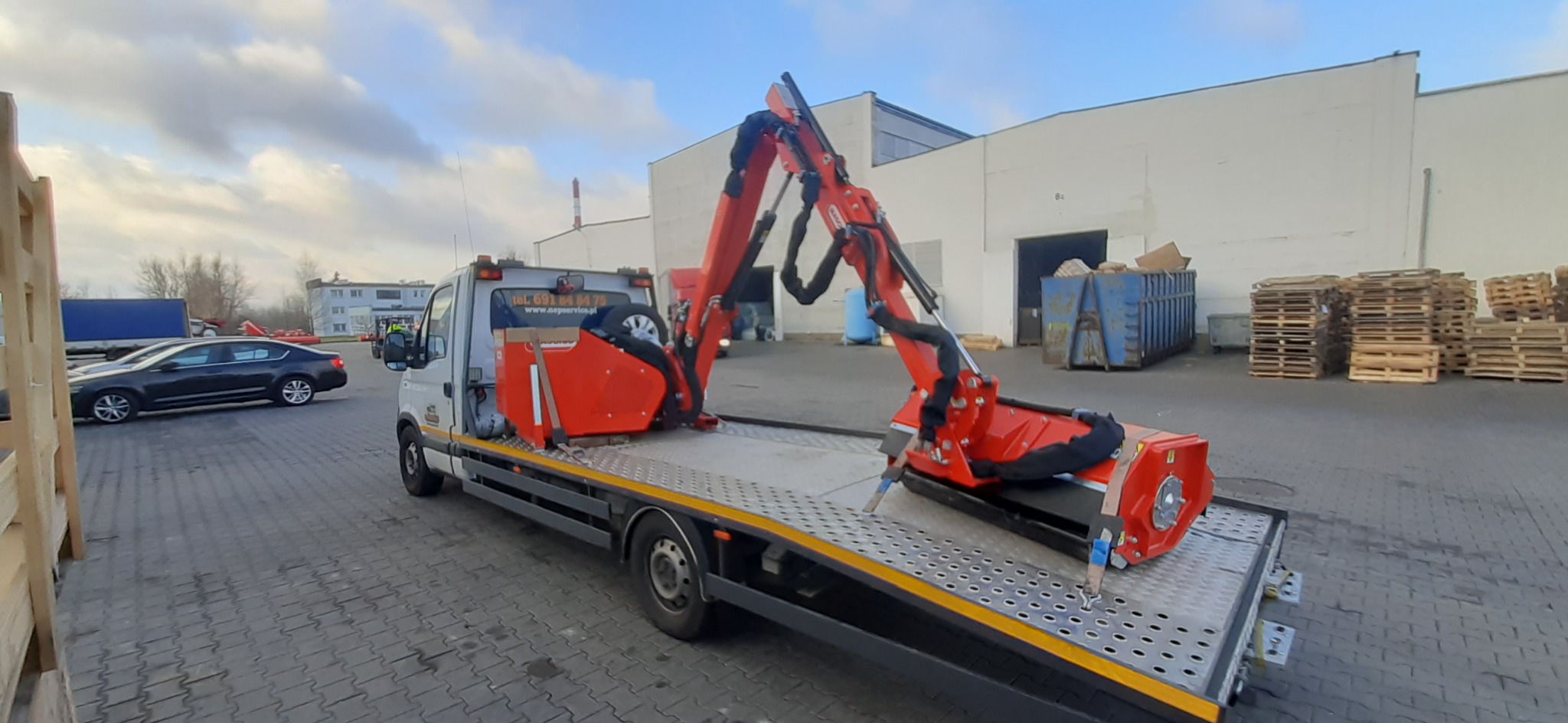 Laweta z zamontowaną czerwoną maszyną rolniczą z wysięgnikiem hydraulicznym na tle magazynu i sterty palet.