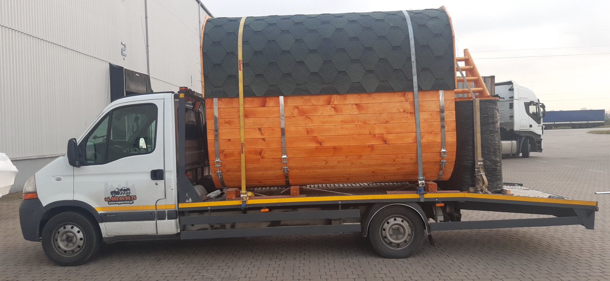 Biała laweta przewozi drewnianą beczkę z dachem pokrytym gontem bitumicznym, zabezpieczoną pasami transportowymi, na tle hali magazynowej i innych pojazdów.