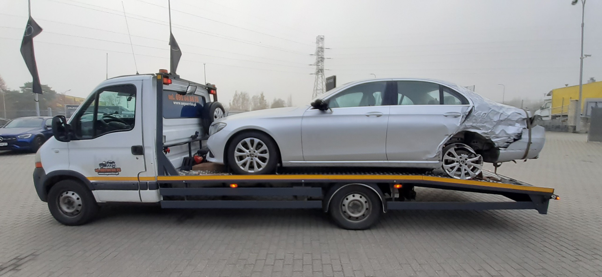 Srebrny samochód osobowy Mercedes z uszkodzonym tyłem załadowany na lawetę, widok z boku, na lawecie widoczne logo firmy.