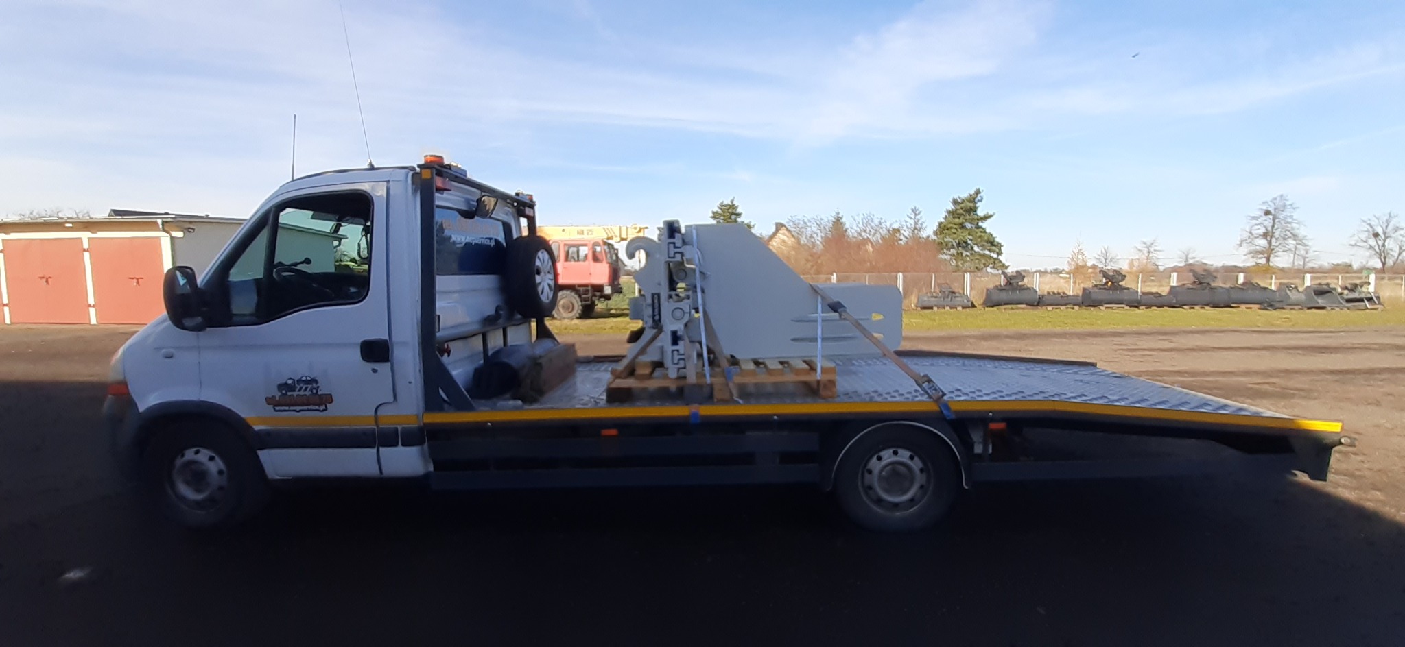 Biała laweta z zabezpieczoną maszyną przemysłową na palecie, z tyłu widoczne inne pojazdy i budynki.