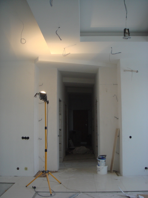Wnętrze w trakcie remontu: widoczne przewody elektryczne wystające ze ściany i sufitu, lampa budowlana na trójnogu, niedokończona podłoga, puszki z farbą i przejście do kolejnego pomieszczenia.