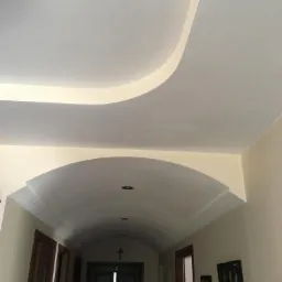 Korytarz z dekoracyjnym sufitem podwieszanym z elementami łukowymi i oświetleniem ambientowym wzdłuż krawędzi, widoczne drzwi i obrazy na ścianie.