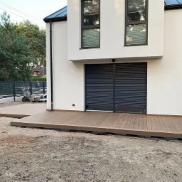 Nowoczesny taras z desek kompozytowych w kolorze brązowym, przylegający do białego budynku z szarymi roletami, widoczny fragment ogrodu z siatką ogrodzeniową i wózkiem narzędziowym.