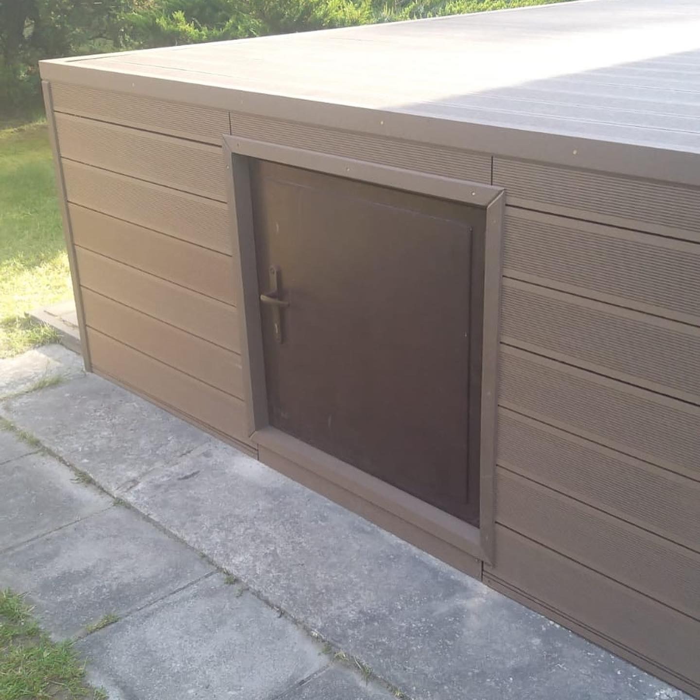 Zamykana skrzynia ogrodowa z brązowych desek kompozytowych, z ciemnymi drzwiczkami i metalowym uchwytem, na betonowym chodniku.